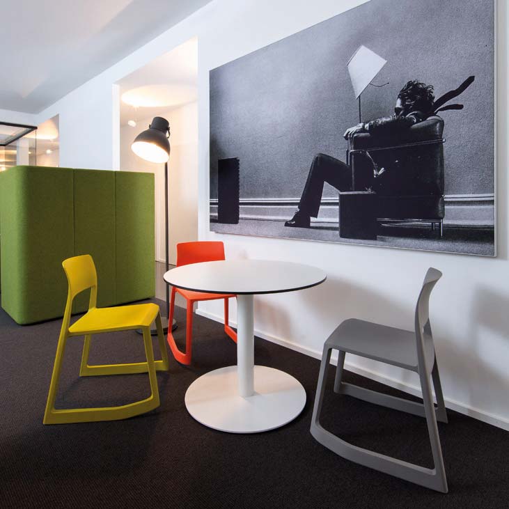 Drei Stühle in unterschiedlicher Farbe stehen um einen kleinen runden Tisch. An der Wand hängt ein großes moderne Gemälde.