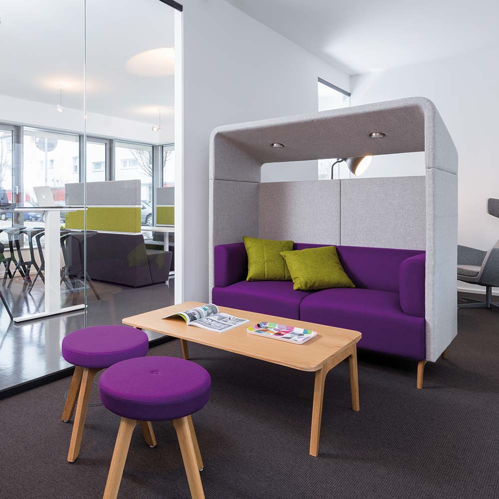 Modernes Büro mit lila Sofa und grau gepolsteter Überdachung hinter einem schmalen Couchtisch aus Holz und zwei lila Hocker.