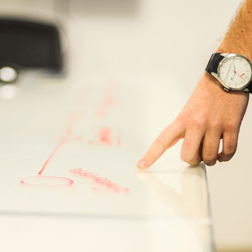 Eine männliche Hand mit Armbanduhr am Gelenk deutet mit dem Zeigefinger auf ein beschriftetes Whiteboard.